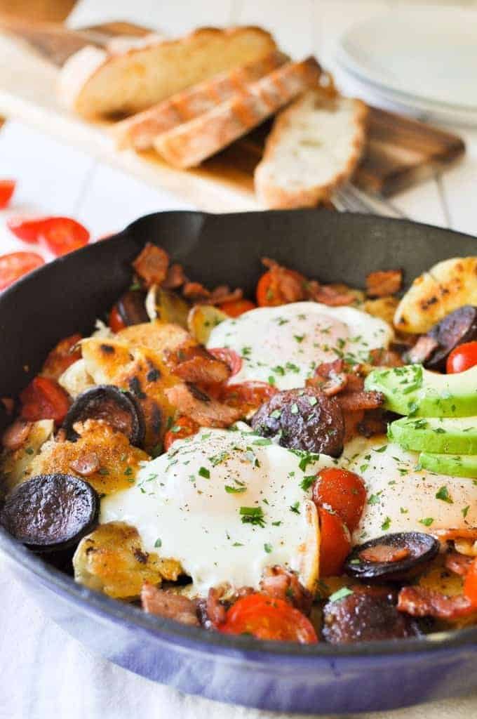 Chorizo Breakfast Hash with crispy smashed potatoes and eggs in a cast iron skillet with avocado on the side with bread in the background.