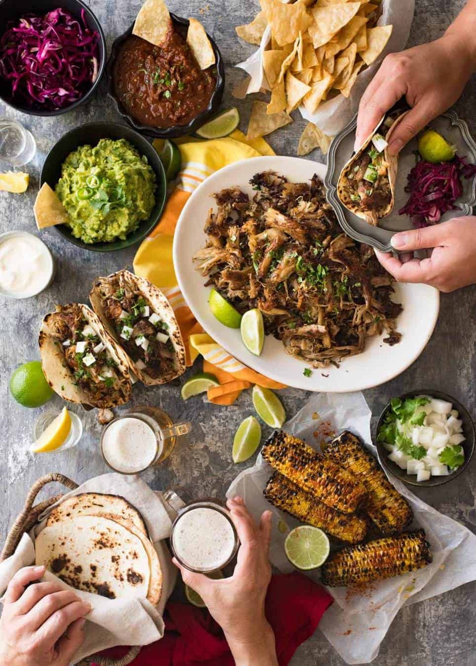 Overhead photo of Mexican Fiesta taco dinner party, with pork carnitas, tacos, grilled corn, guacamole, salsa, pickled red cabbage, tortilla chips and taco sides.