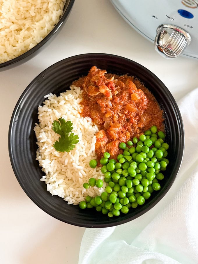 Slow Cooker Butter Chicken served in a black bowl with a serving a white rice, green peas and garnished with a sprig of fresh coriander.